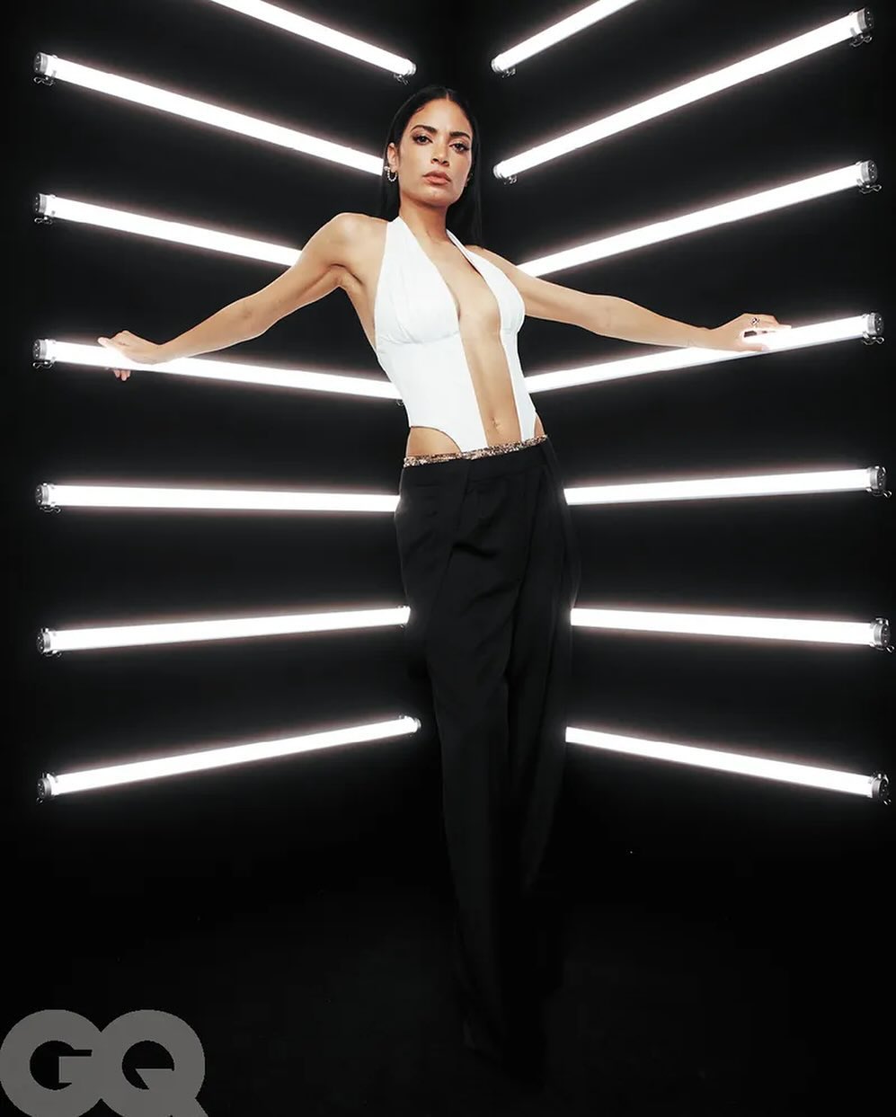 A stylish adult woman with dark hair and a white halter top poses in front of horizontal neon lights against a dark background.
