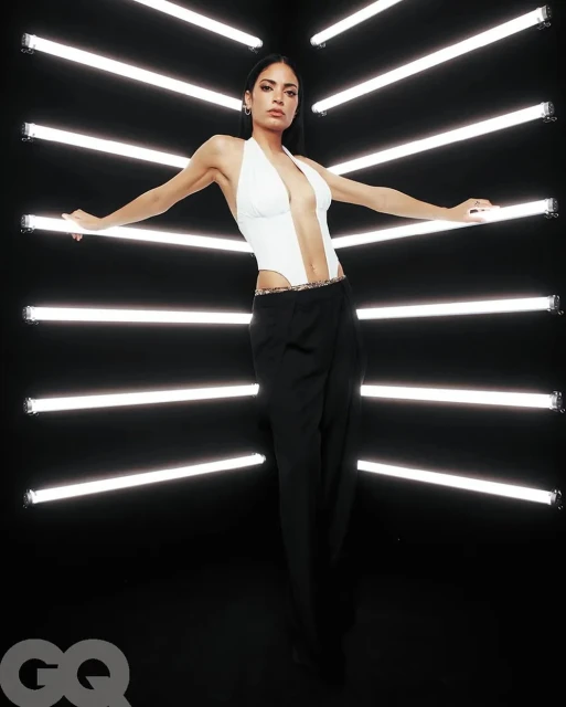A stylish adult woman with dark hair and a white halter top poses in front of horizontal neon lights against a dark background.
