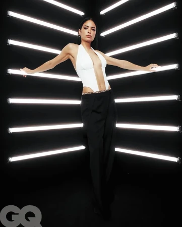 A stylish adult woman with dark hair and a white halter top poses in front of horizontal neon lights against a dark background.