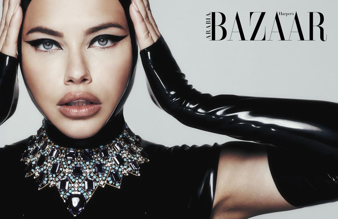 Close-up portrait of an adult female fashion model wearing a black latex hood and a large jeweled necklace, with Harper's Bazaar logo.