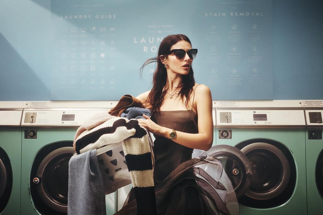 A woman in sunglasses holding a pile of laundry in a laundromat, wearing a dress and watch with washing machines in the background.