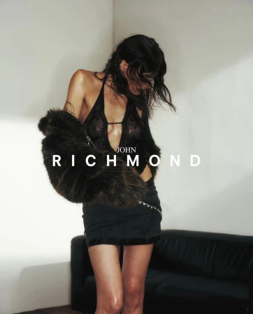Woman in sheer black halter top holding a furry brown jacket standing before a dark sofa
