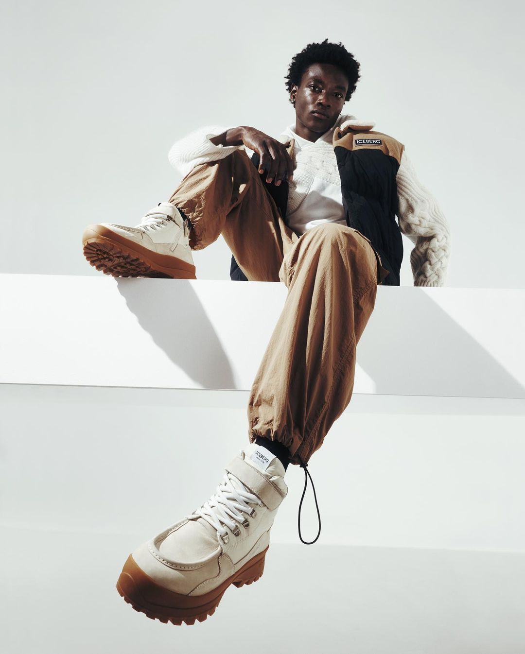 An adult Black male sits on a white ledge wearing beige pants, a white sweater, and white boots in a studio setting with a white backdrop.