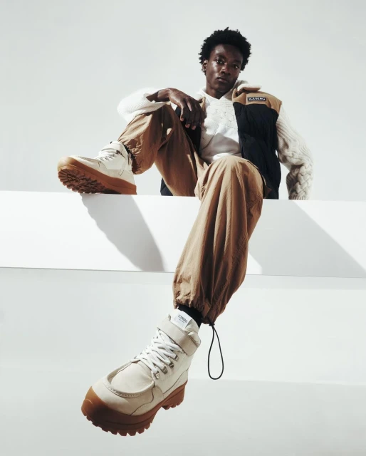 An adult Black male sits on a white ledge wearing beige pants, a white sweater, and white boots in a studio setting with a white backdrop.