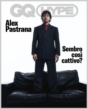 Alex Pastrana standing on a black leather sofa against a white background, wearing a dark double-breasted suit and red shirt.