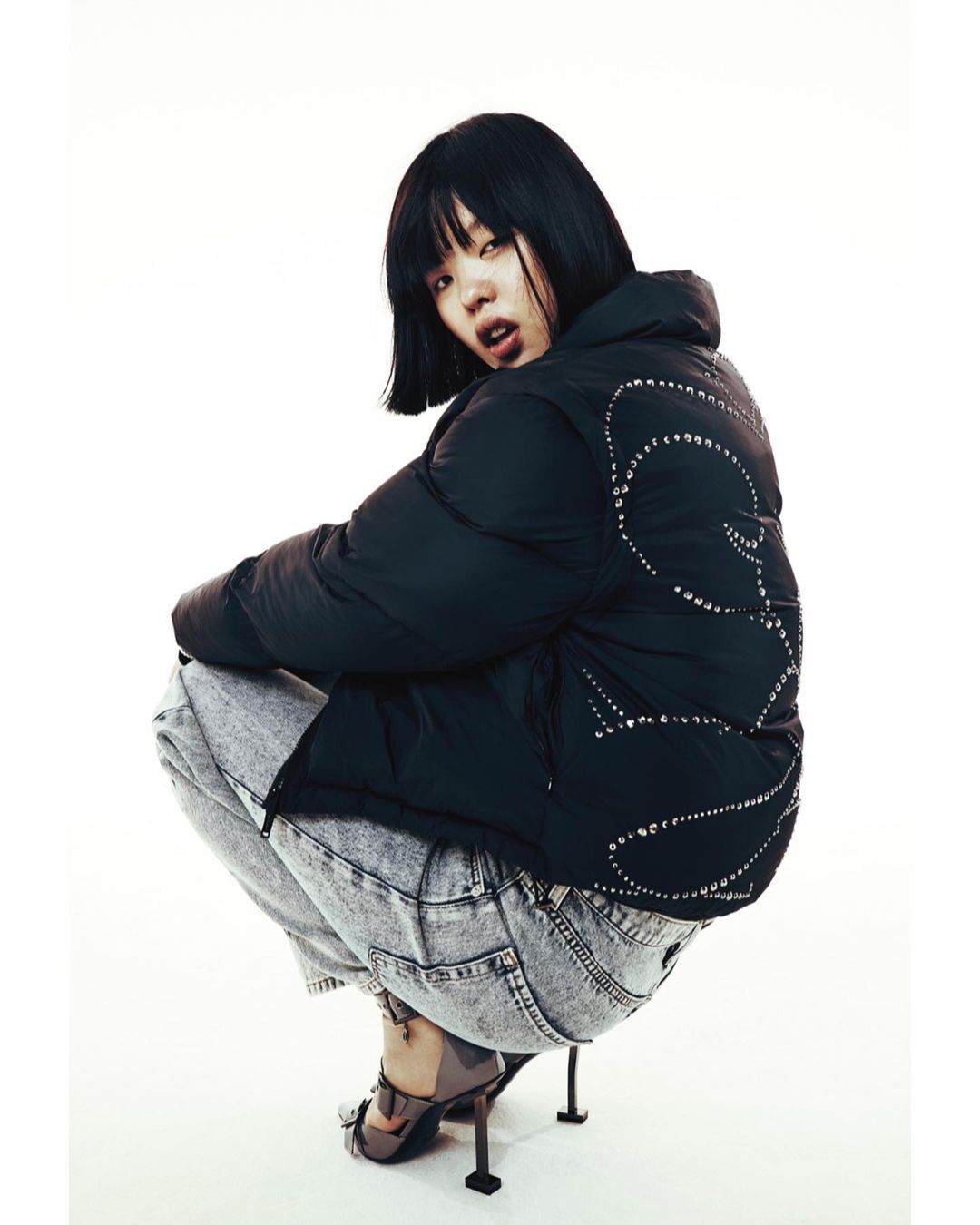 Stylish woman in black puffer jacket and acid wash jeans crouching indoors, posing against a white backdrop in a fashion editorial shot.