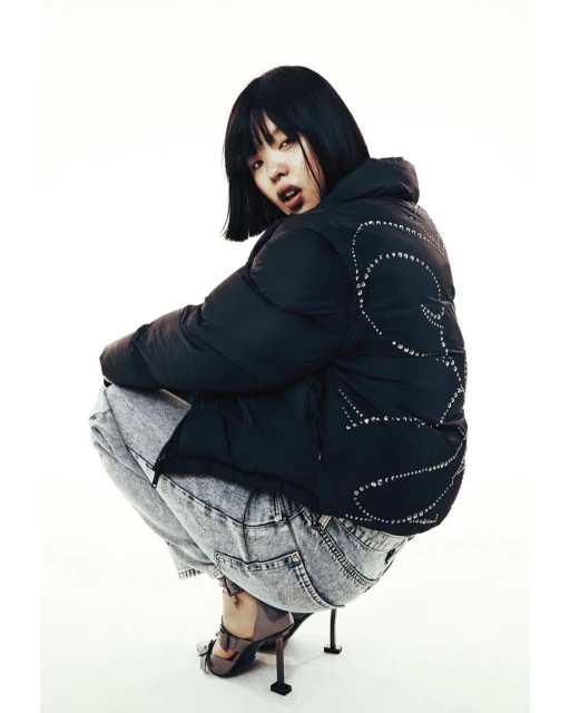 Stylish woman in black puffer jacket and acid wash jeans crouching indoors, posing against a white backdrop in a fashion editorial shot.