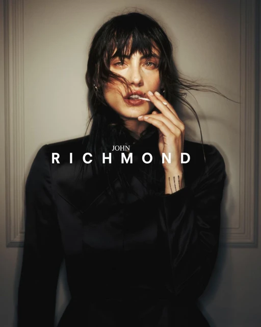 Portrait of woman in black top holding lollipop, featuring 'John Richmond' text. Fashion shot with subdued lighting, neutral background.