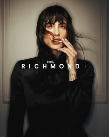 Portrait of woman in black top holding lollipop, featuring 'John Richmond' text. Fashion shot with subdued lighting, neutral background.