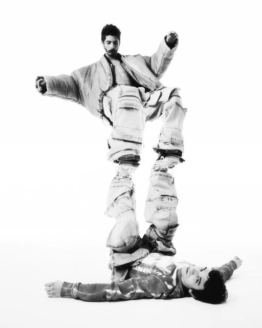 Artistic black and white studio shot of two men in unique textured clothing, one balanced atop the other, showcasing a creative fashion concept.