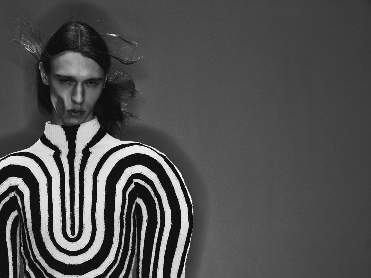 Black and white portrait of a person with long hair wearing a striped sweater, with soft studio lighting and a neutral background.