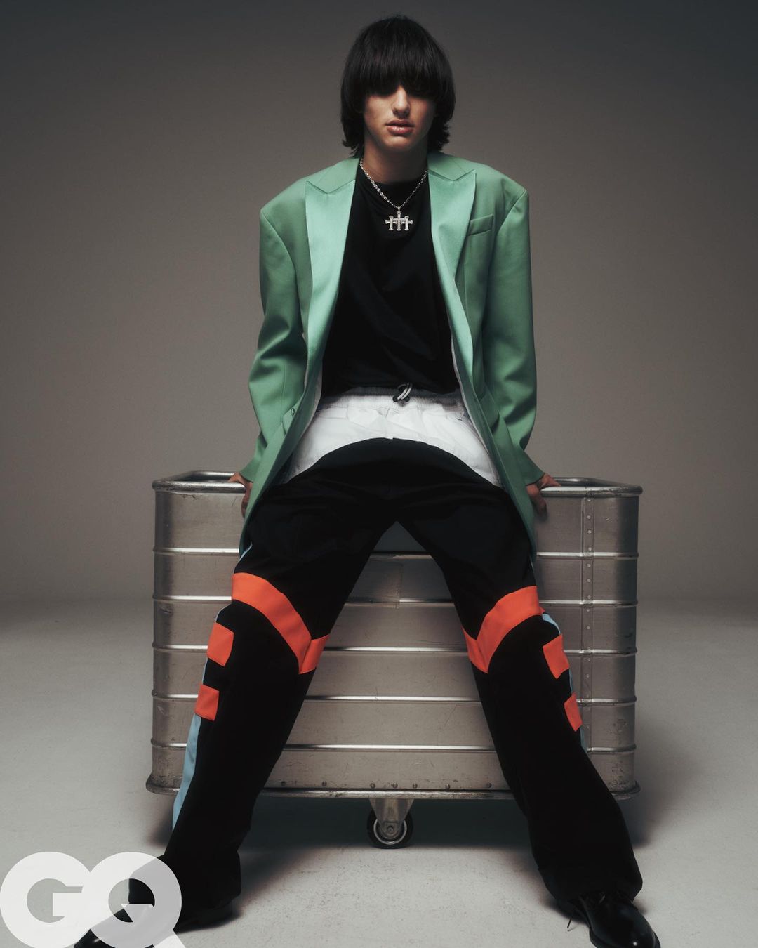 A young adult poses in a fashion editorial wearing an oversized blazer and track pants while sitting on an aluminum trunk.