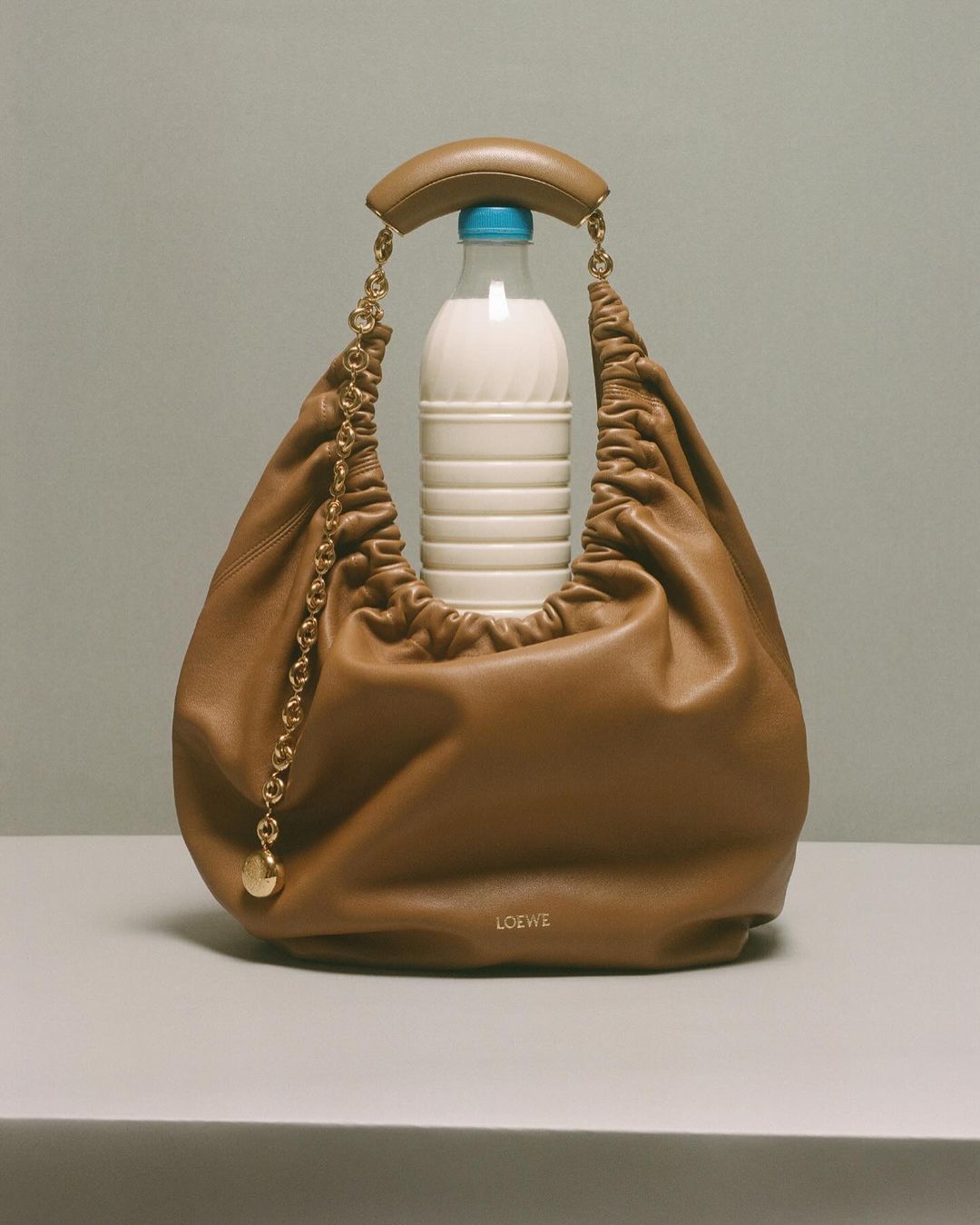 Studio shot of a tan leather handbag with gold chain detail, styled with a blue and white bottle. Neutral background and soft lighting.