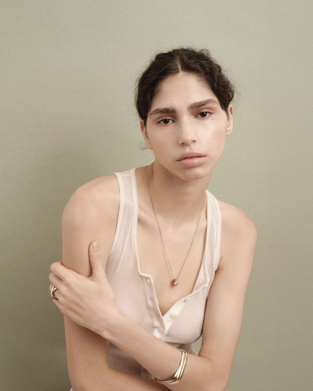 Young woman portrait in white tank top against a solid background. She wears gold jewelry and poses in a studio with neutral lighting.