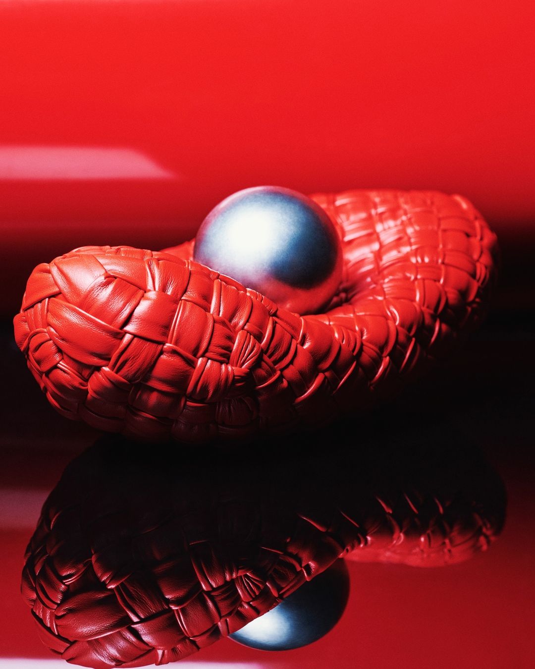 A metallic ball rests in a woven red leather container, set against a reflective red background in a studio setting.