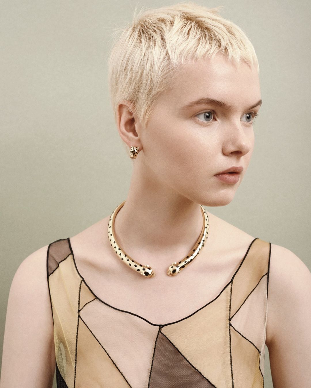Woman with a short blonde haircut wears a gold-tone cheetah-head collar necklace and earrings indoors against a plain backdrop.