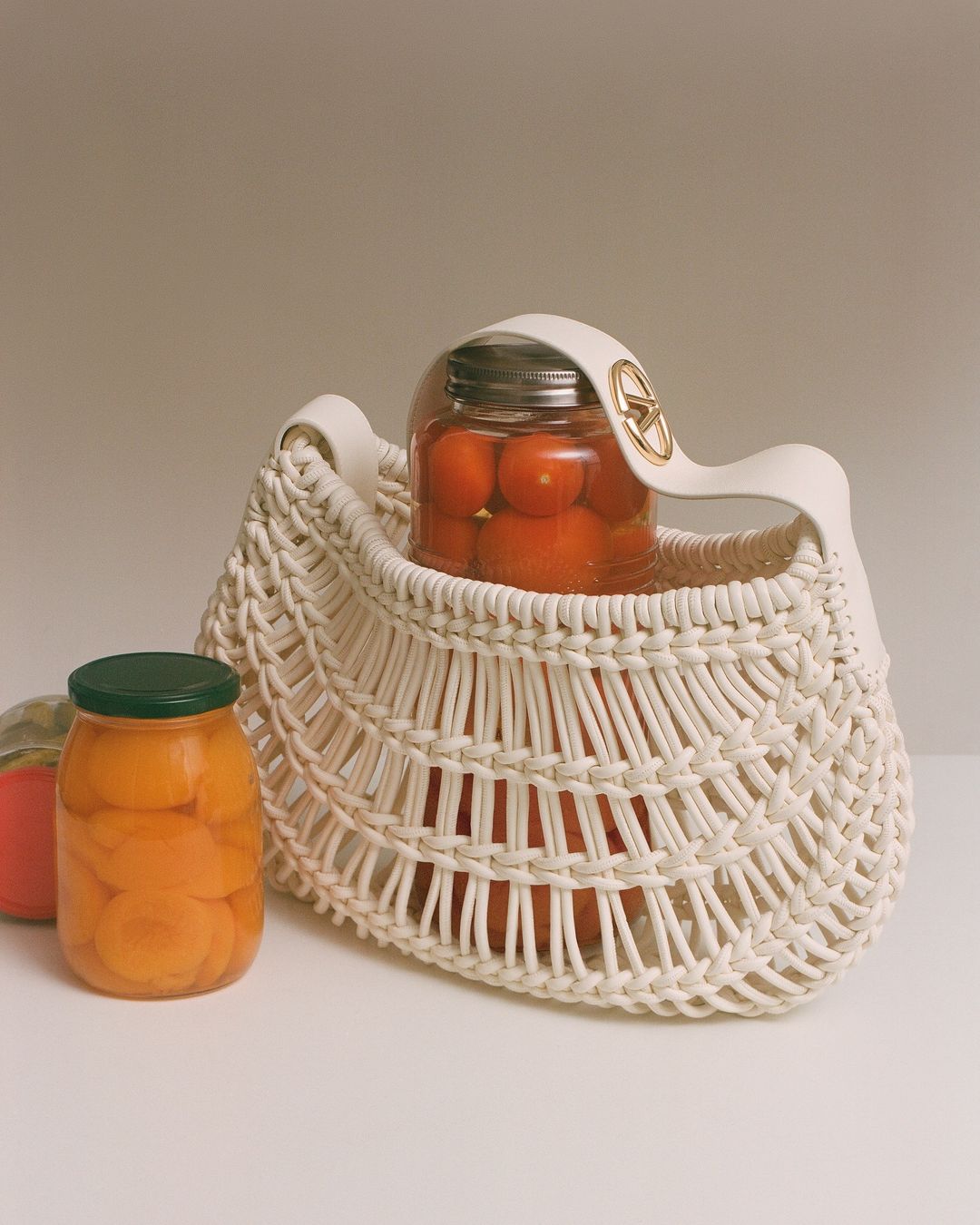 Wicker handbag containing a jar of tomatoes, beside a jar of canned peaches on a white surface against a neutral backdrop.