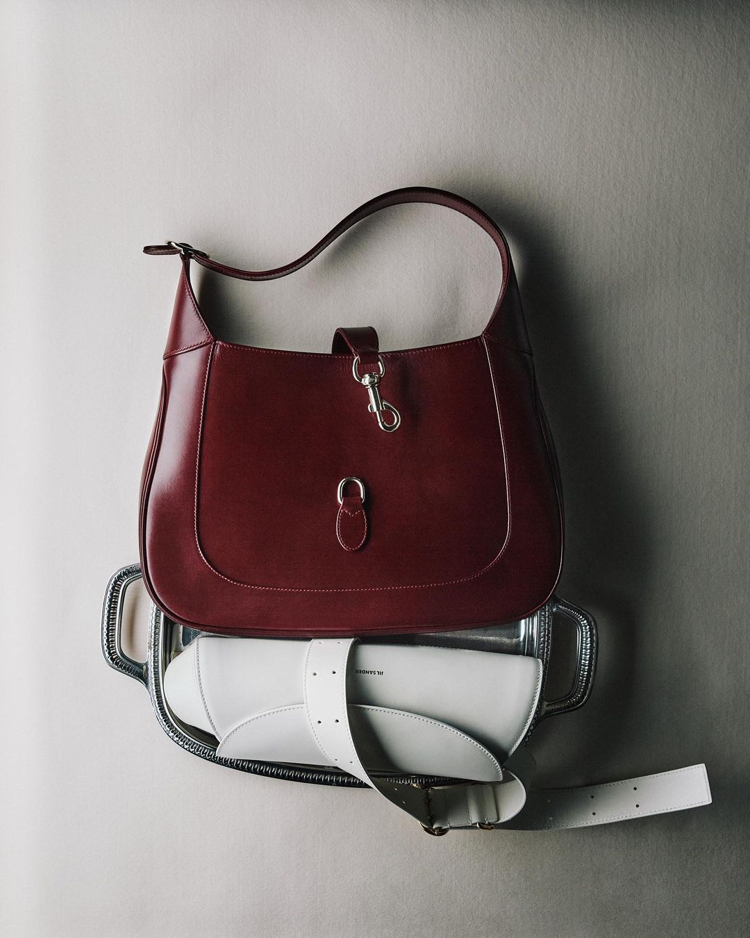 Elevated product shot of a burgundy leather hobo bag atop a silver tray, with a white purse and strap, against a neutral backdrop.