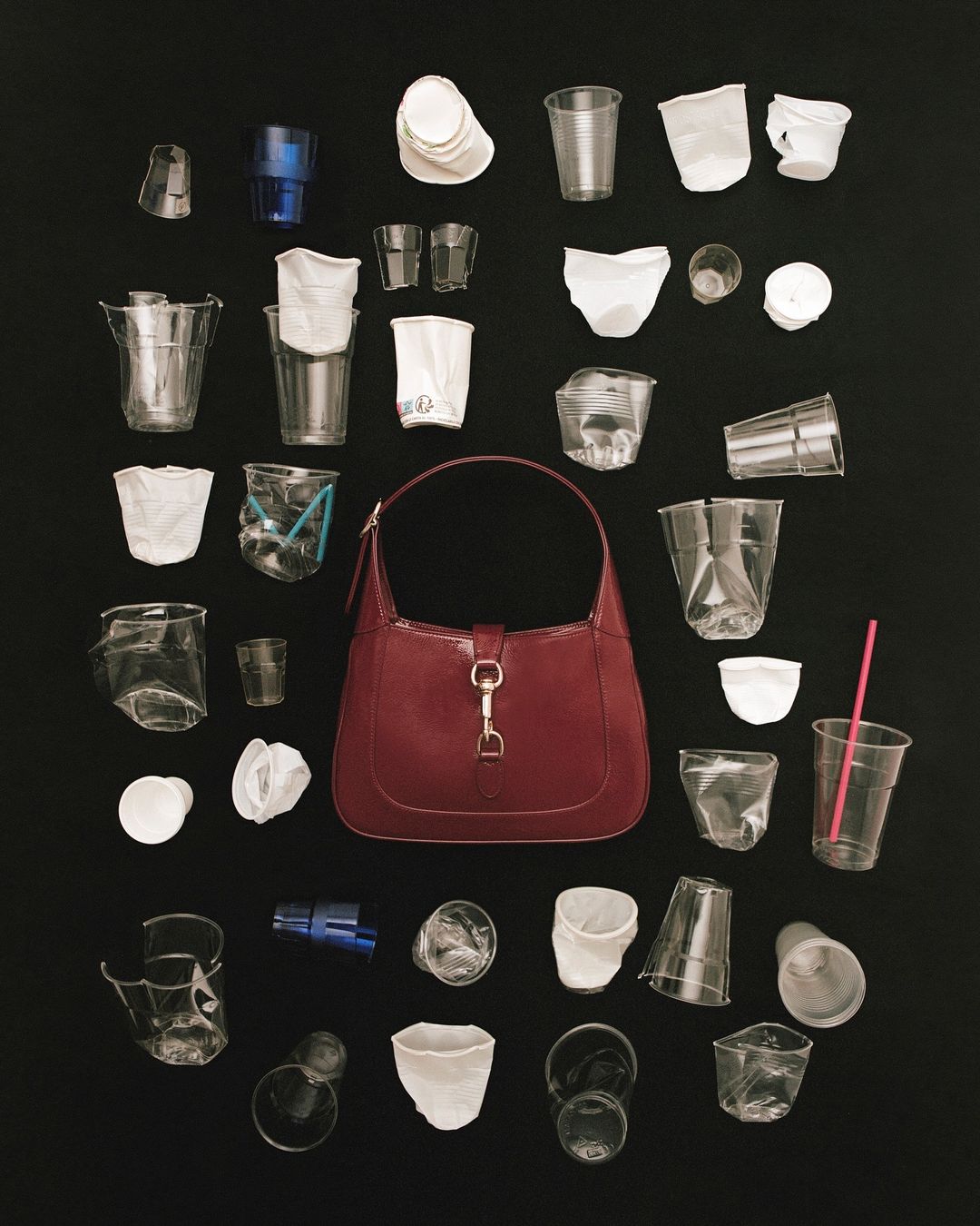 Overhead shot of a dark red purse surrounded by assorted used plastic cups on a black background, highlighting waste and fashion.
