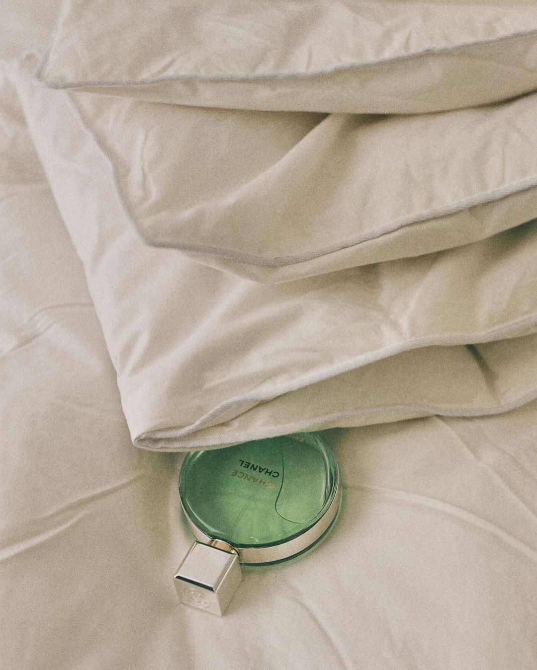 Overhead shot of a green Chanel perfume bottle on a white quilted duvet, set against a background of folded duvet layers.