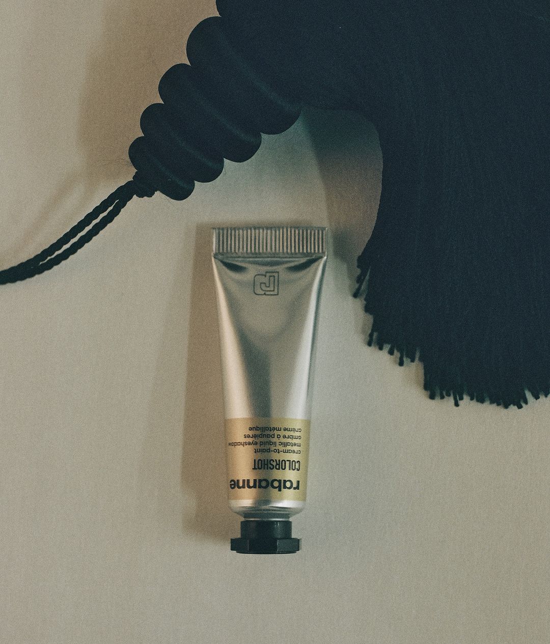 Silver tube of Rabanne Colorshot cosmetic product against a cream wall with a black tassel detail in the upper right corner.