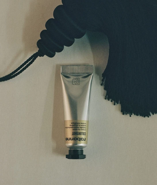 Silver tube of Rabanne Colorshot cosmetic product against a cream wall with a black tassel detail in the upper right corner.