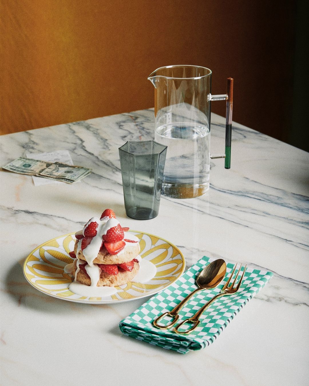 Strawberry shortcake dessert with cream on a patterned plate, next to a pitcher of water and a money stack on a marble table.