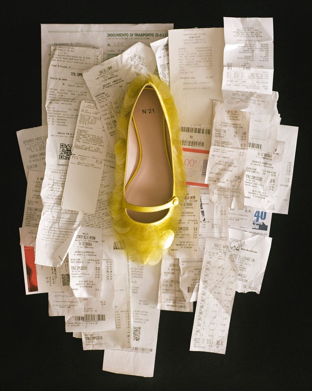 High-angle shot: a yellow ballet flat with a strap is displayed on a pile of financial receipts against a solid black background.