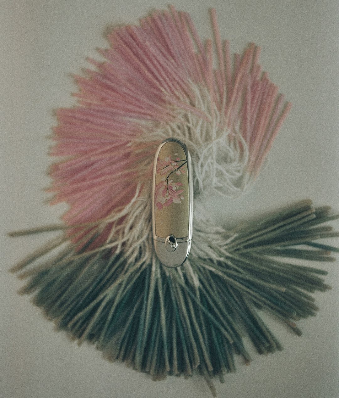 Cosmetic tube with silver accents and floral details surrounded by pink, white, and teal fringe on a white background.