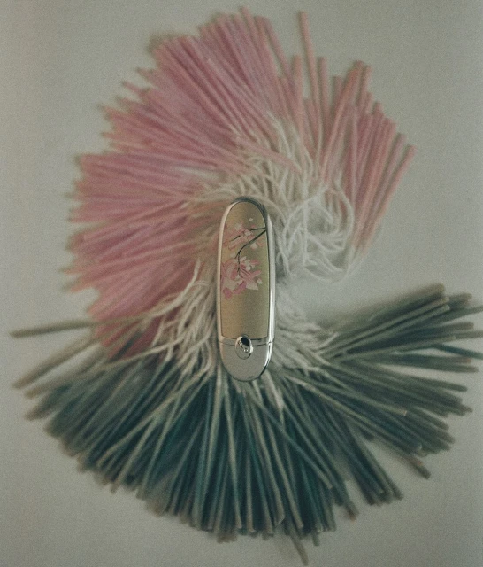 Cosmetic tube with silver accents and floral details surrounded by pink, white, and teal fringe on a white background.