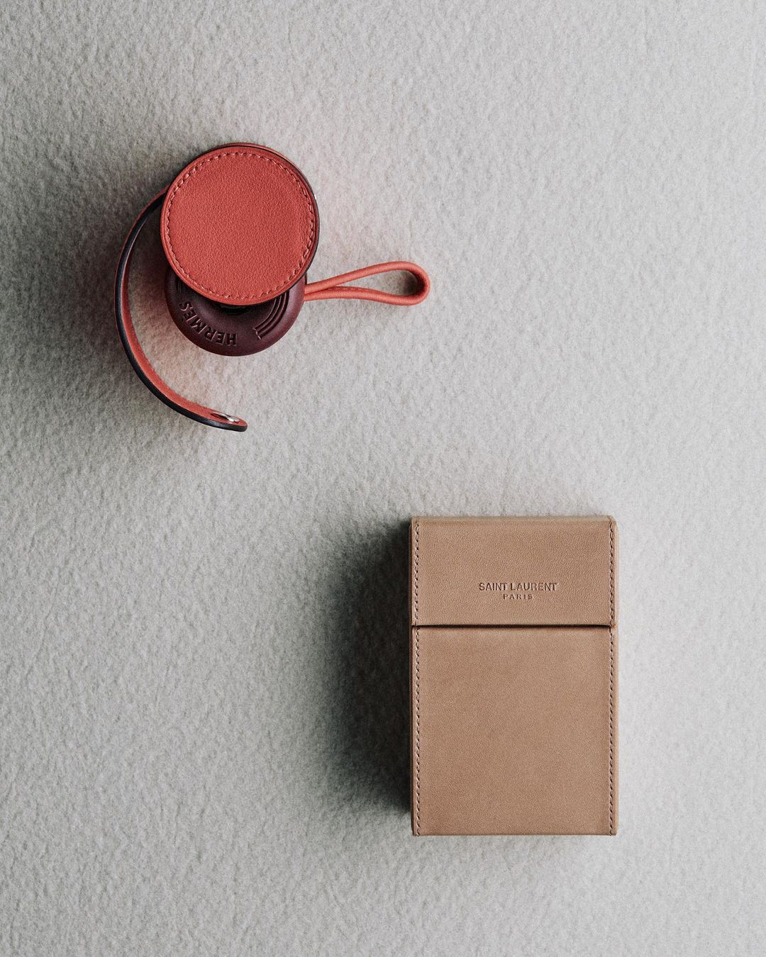 Elevated view of a coral Hermes object and a tan Saint Laurent box on a textured grey surface, emphasizing luxury fashion accessories in a studio setting.