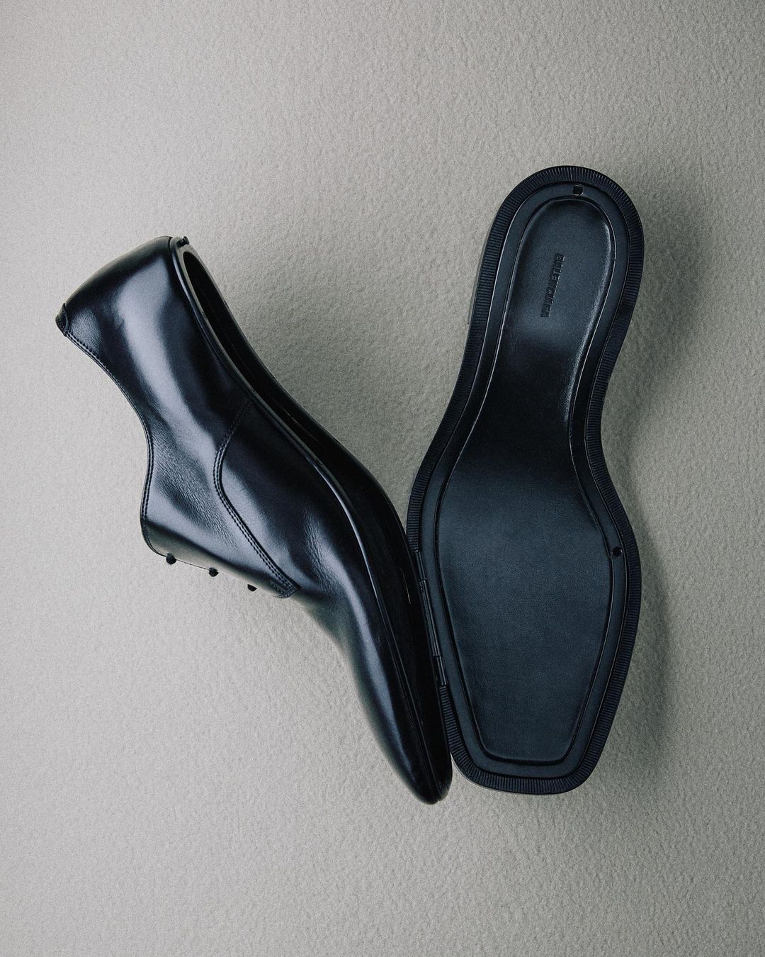 Overhead shot of a pair of black leather oxford shoes, one showing the upper and one showing the sole on a pale grey textured background.