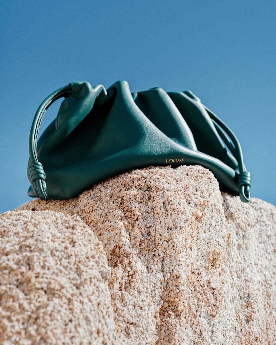 Teal Loewe clutch handbag on a textured rock formation against a clear blue sky. Fashion still life, studio lighting.