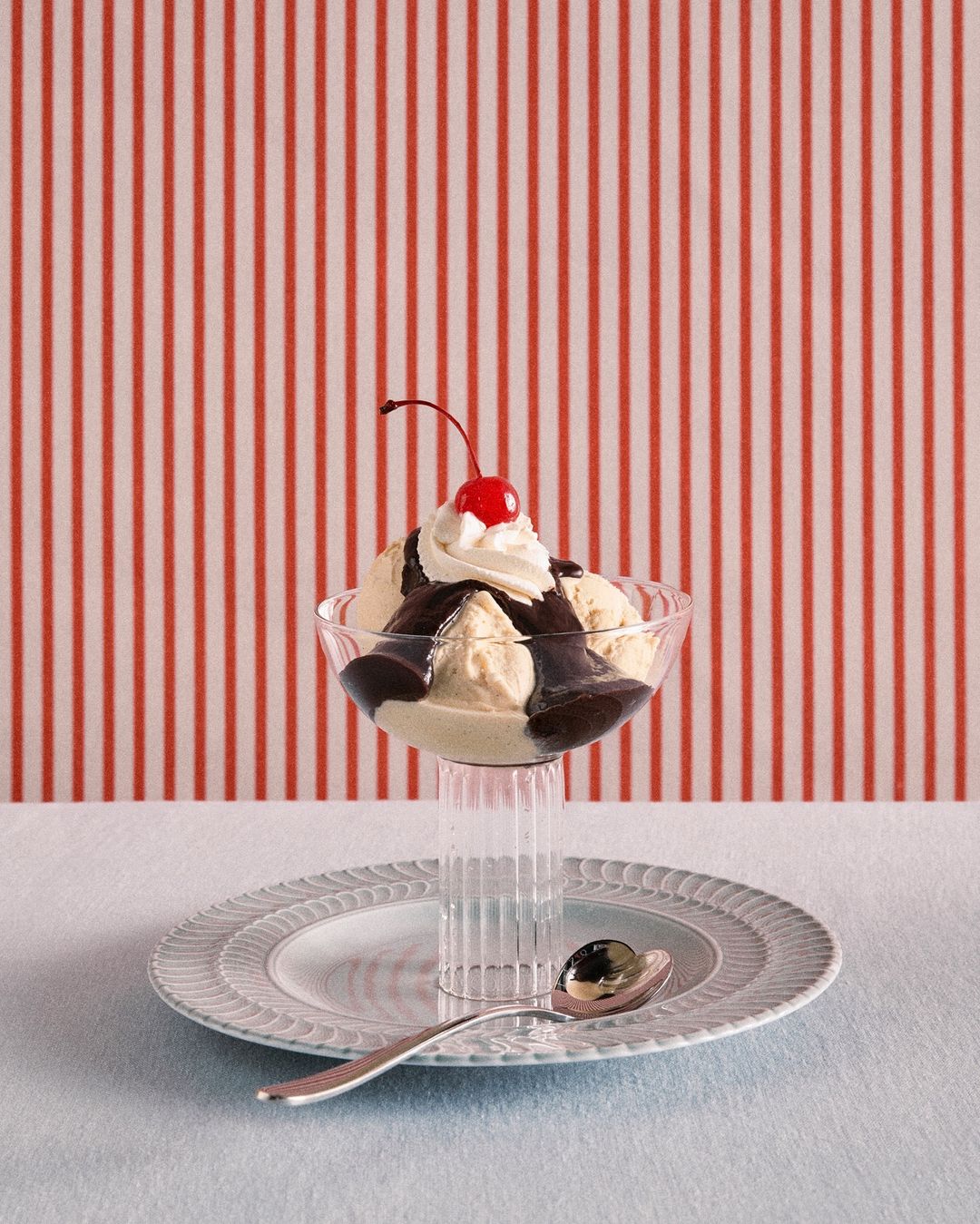 A glass sundae dish filled with ice cream, chocolate sauce, whipped cream, and a cherry on top, resting on a plate with a spoon.