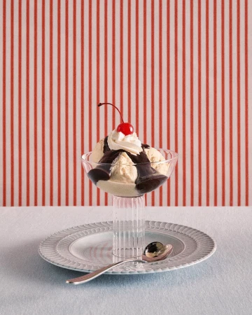 A glass sundae dish filled with ice cream, chocolate sauce, whipped cream, and a cherry on top, resting on a plate with a spoon.