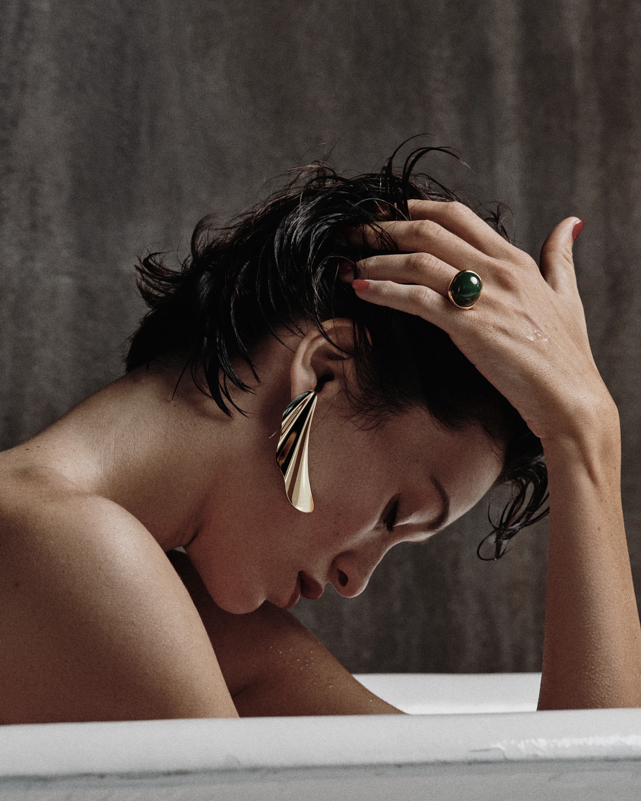 Close-up portrait of a woman with wet dark hair in a bathtub wearing large gold statement earrings and a green gemstone ring.
