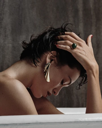 Close-up portrait of a woman with wet dark hair in a bathtub wearing large gold statement earrings and a green gemstone ring.