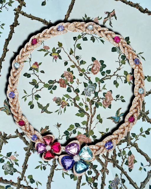Braided gold necklace with diamonds and colorful floral gemstone accents on a botanical wallpaper backdrop, jewelry product shot.