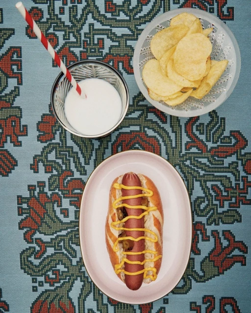 Overhead shot of a hot dog with mustard, milk in a glass with a straw, and potato chips on a patterned blue tablecloth.