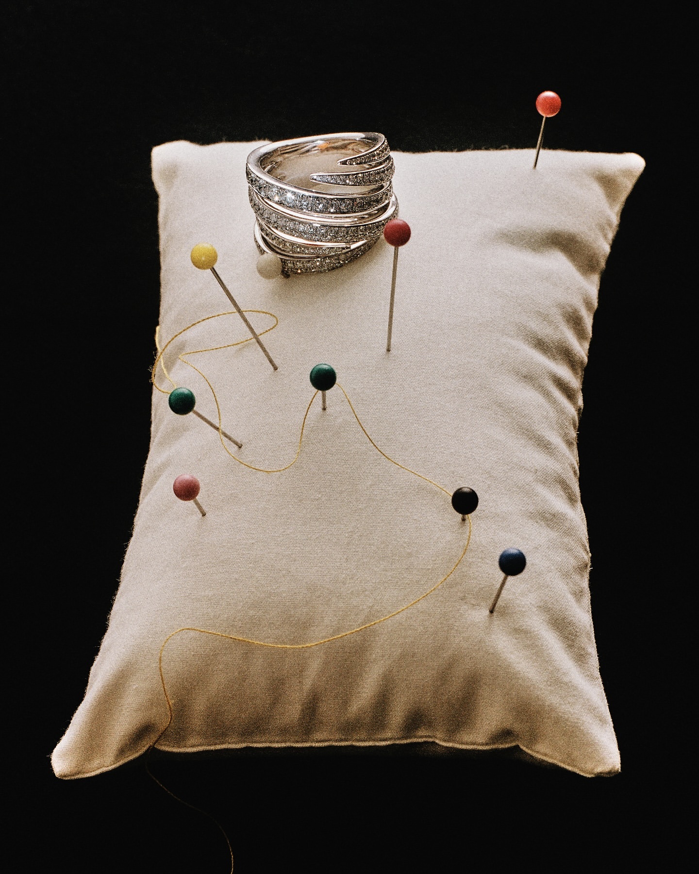 Close-up of a silver diamond ring on a beige pin cushion with multicolored pins and yellow thread on a black background.