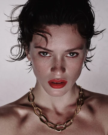 Close-up portrait of a woman with wet dark hair, bold red lipstick, and a gold chain necklace against a light background.