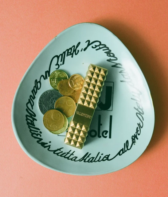 Overhead shot of a Valentino lipstick, euro coins, and a light-blue dish with the word 'Hotel' on a pink background, studio lighting.