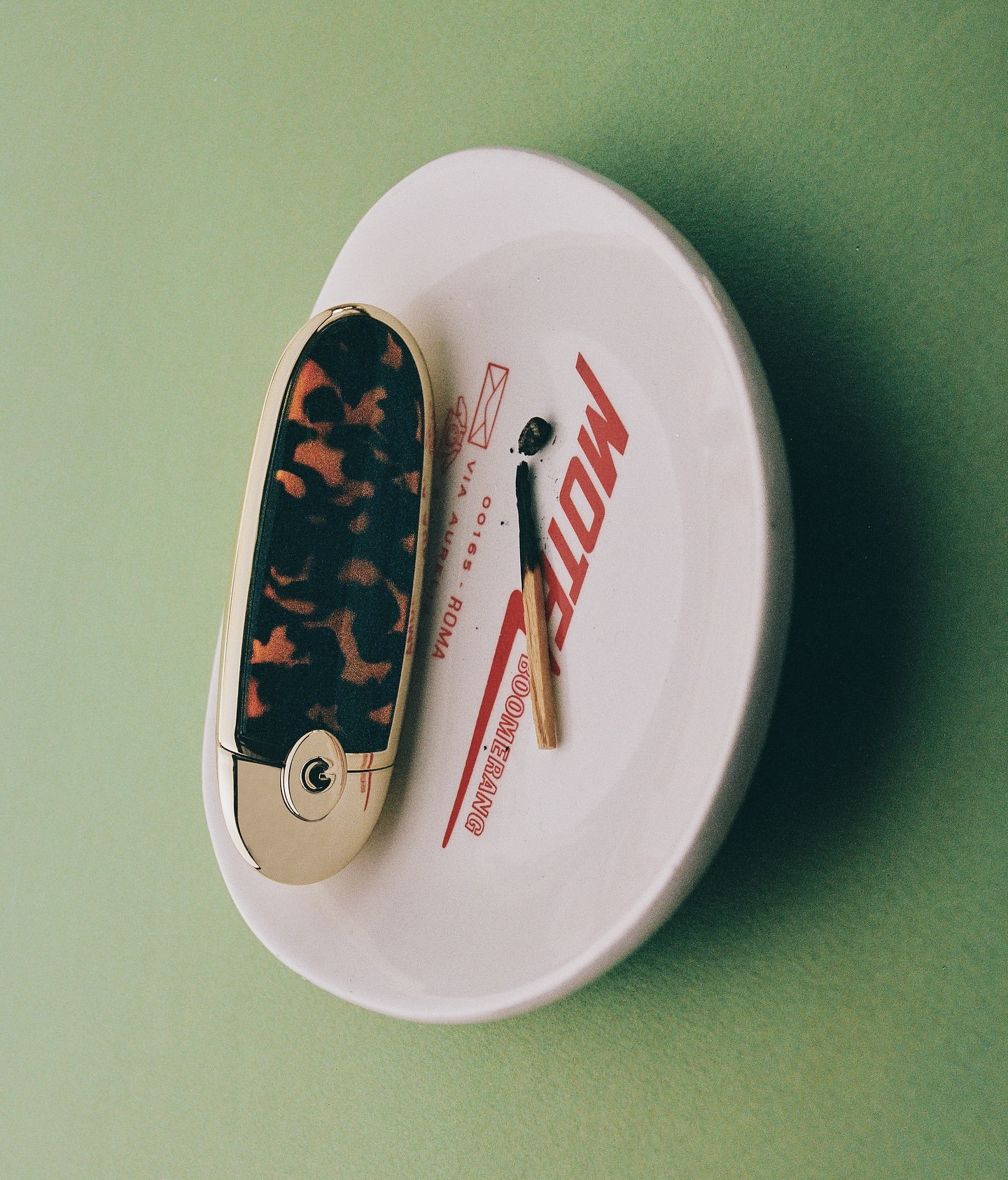 Close-up of a vintage tortoiseshell lipstick case and a spent matchstick on a small white dish with red 'Motel' text, against a green backdrop.