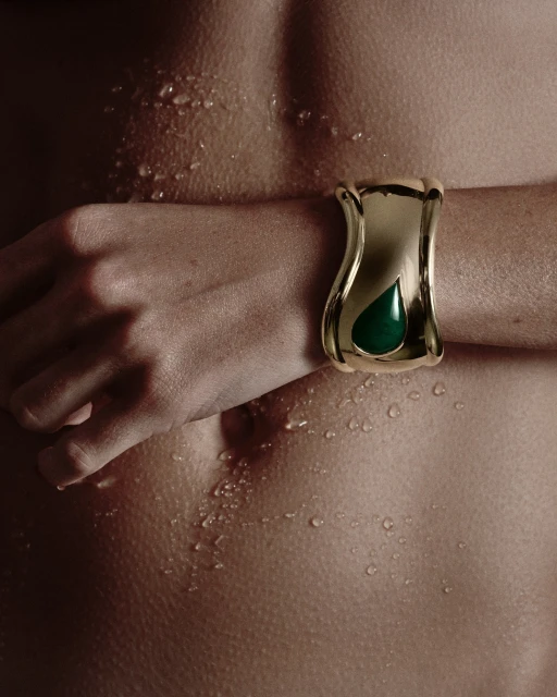 Close-up of a human torso and arm, adorned with a gold and green cuff bracelet, accented by water droplets across the skin.