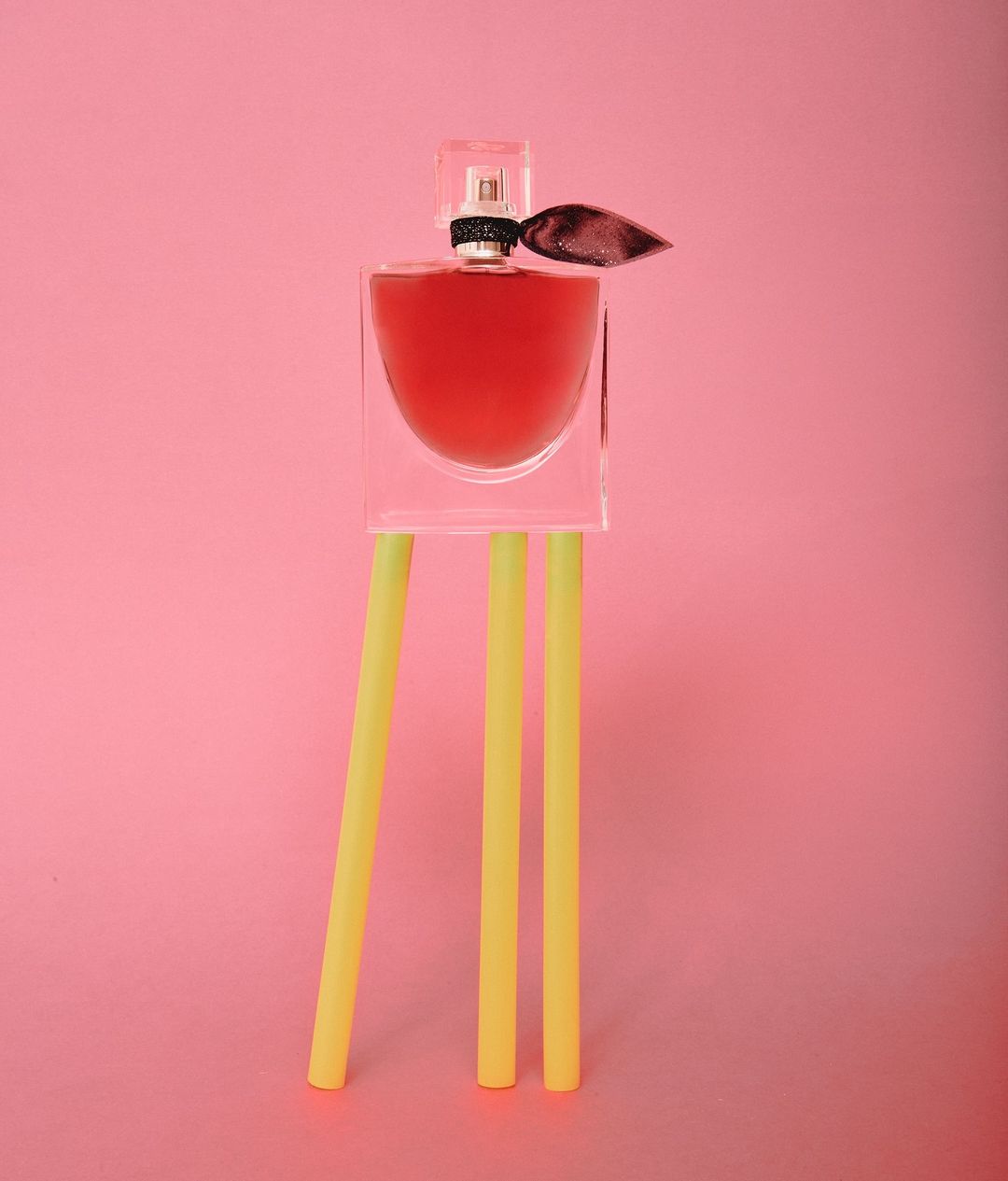 Perfume bottle stands on yellow cylindrical props against a pink background. The bottle has a clear cap and a dark ribbon accent.