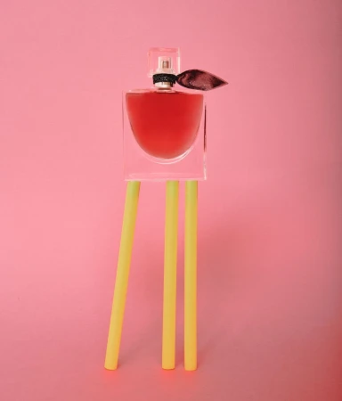 Perfume bottle stands on yellow cylindrical props against a pink background. The bottle has a clear cap and a dark ribbon accent.