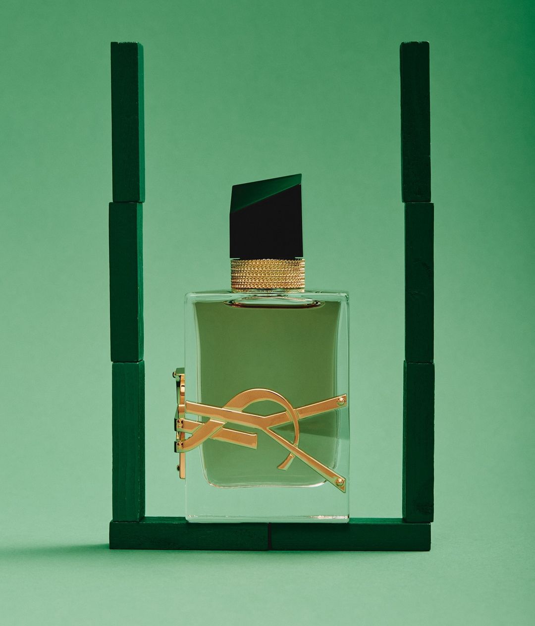 Elevated, studio shot of Yves Saint Laurent cologne bottle against green backdrop, framed by dark green wooden blocks, evoking luxury.