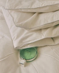 Round green perfume bottle tucked neatly under stacked off-white pillows on a textured bed sheet by Mattia Iotti.