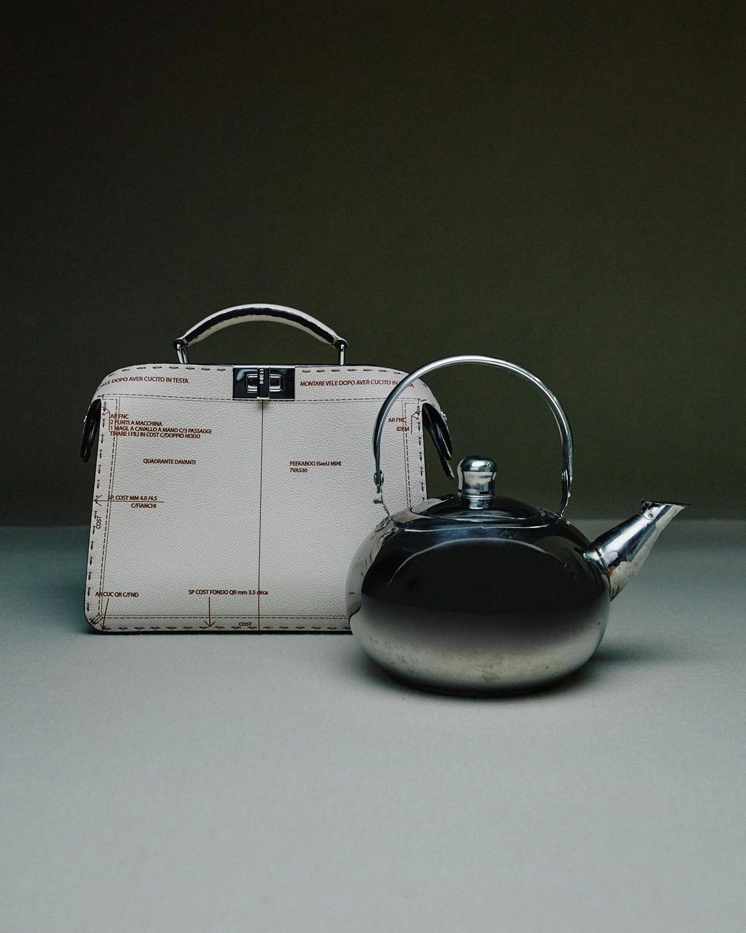 A cream leather structured handbag with printed diagram markings next to a reflective metallic kettle on a grey table against a dark olive background.