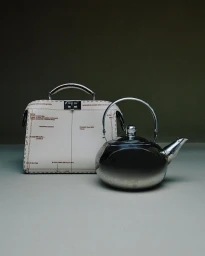 A cream leather structured handbag with printed diagram markings next to a reflective metallic kettle on a grey table against a dark olive background.